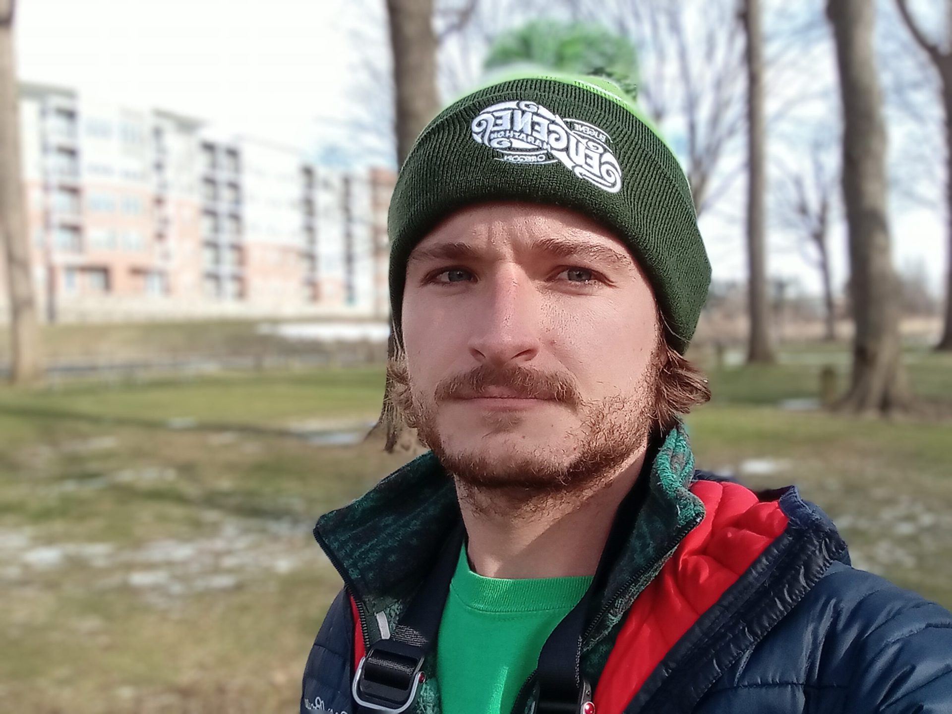 Galaxy A03S肖像Selfie户外一个男人穿着绿色帽子，绿色T恤和红色和黑色外套，与建筑物和树在他身后可见。