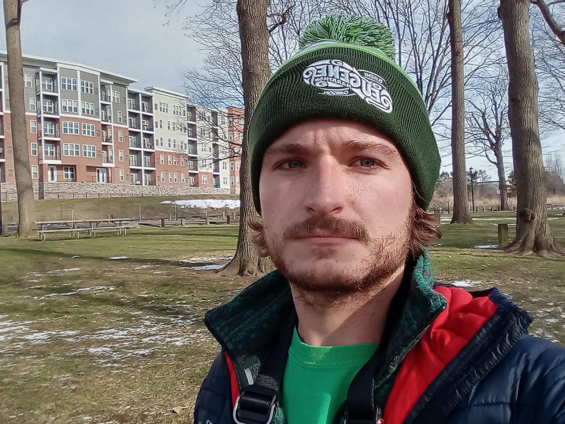 Galaxy A03S标准Selfie在户外的一个男人，带有绿色帽子，绿色T恤和红色和黑色外套，建筑物和树木在他身后可见。